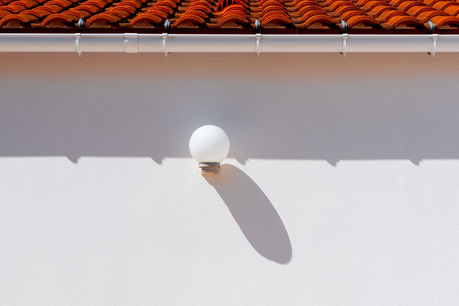 Red clay roof tiles at the eaves above a white gutter on brackets, over a white rendered wall with a globe wall light