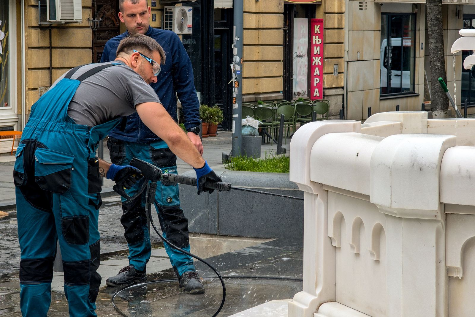 A worker in safety glasses aims a pressure washer lance at the base of a white stone fountain while a colleague watches