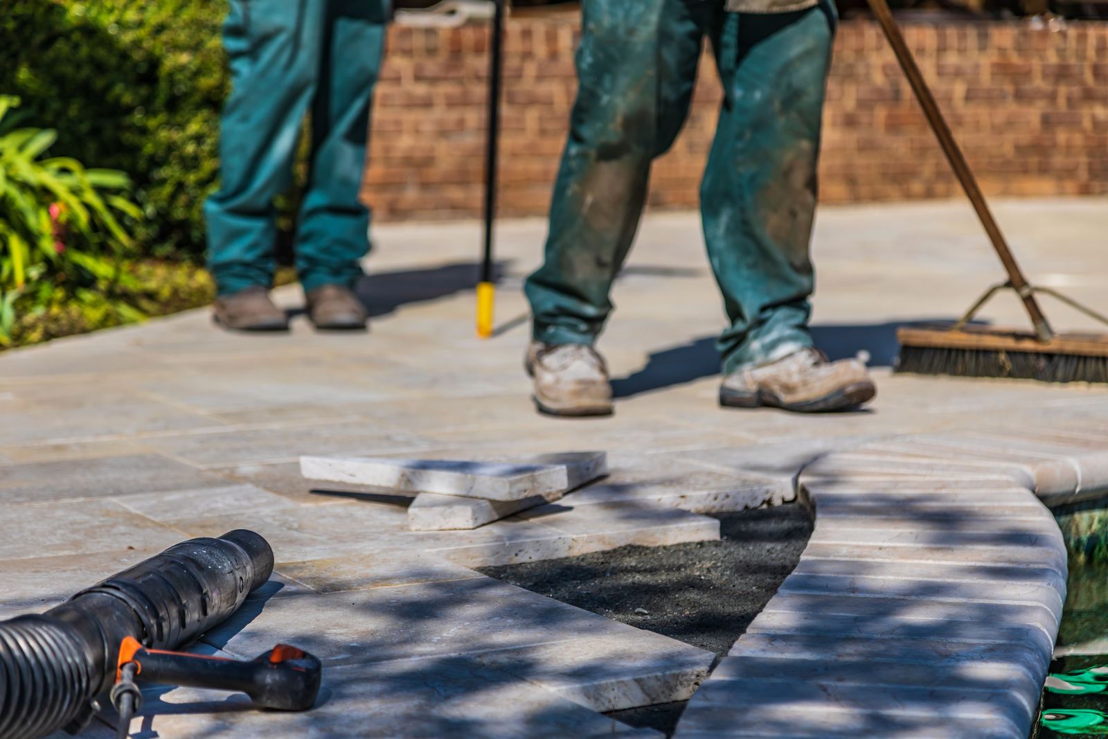 Two workers laying pale stone paving beside a pool, with a broom and a leaf blower