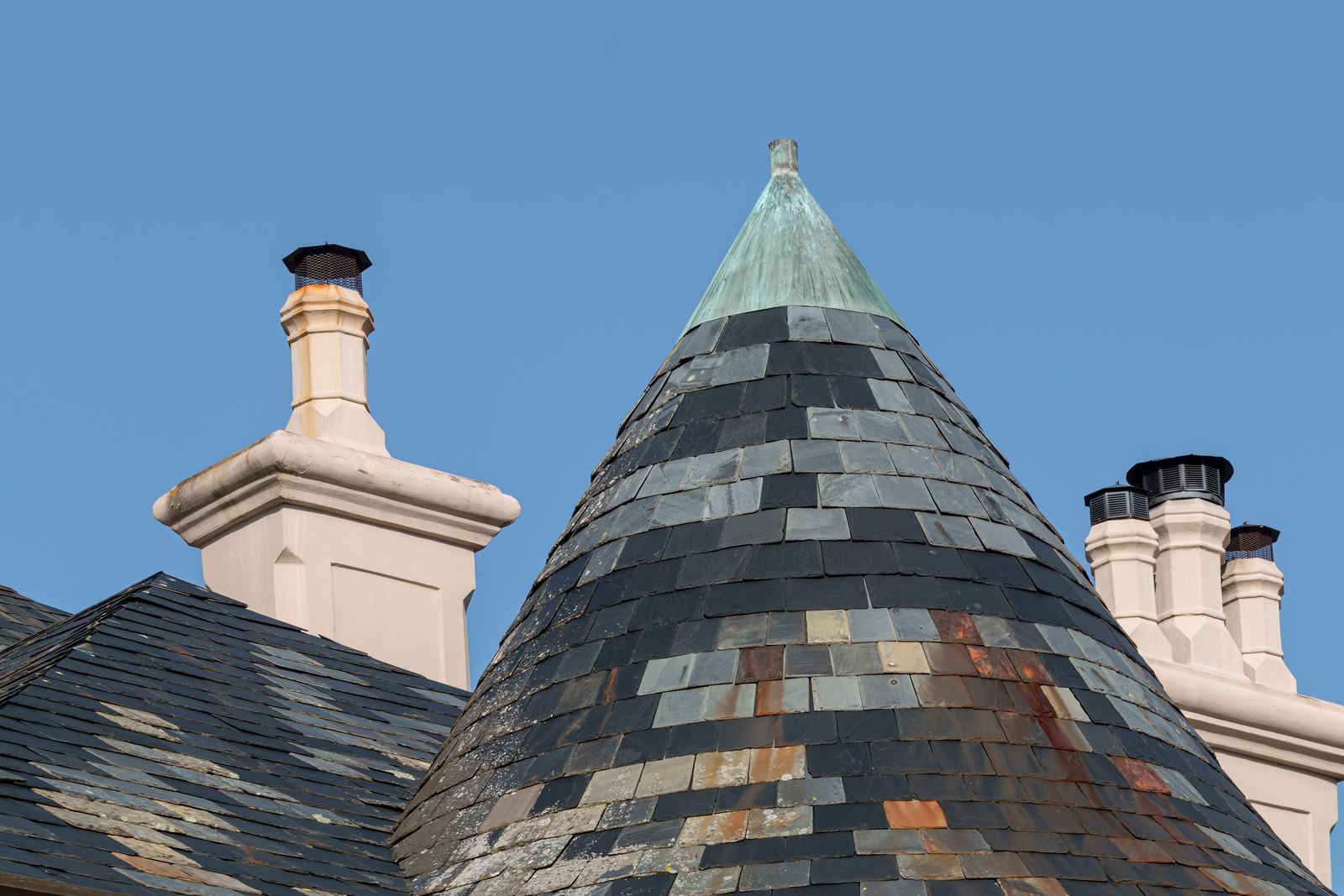 Conical slate turret roof with a green copper finial beside cream chimney stacks under blue sky