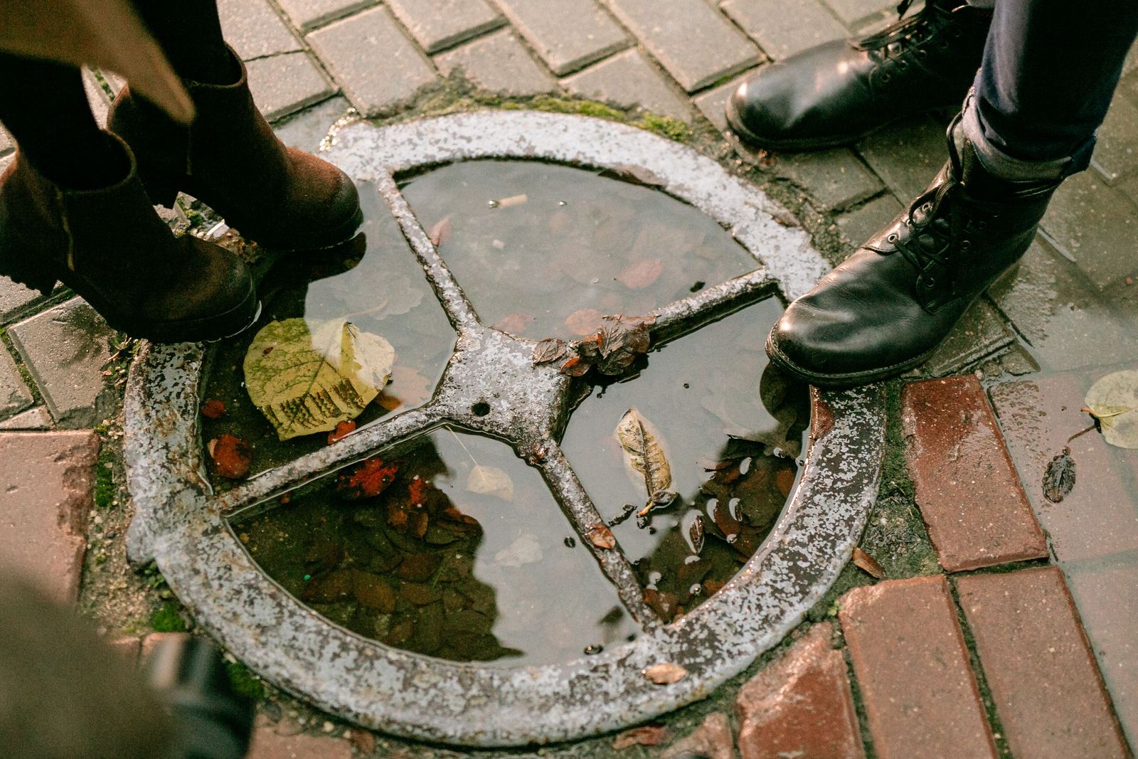 Rainwater pooled in a round drain cover set in brick paving, with people's boots around it