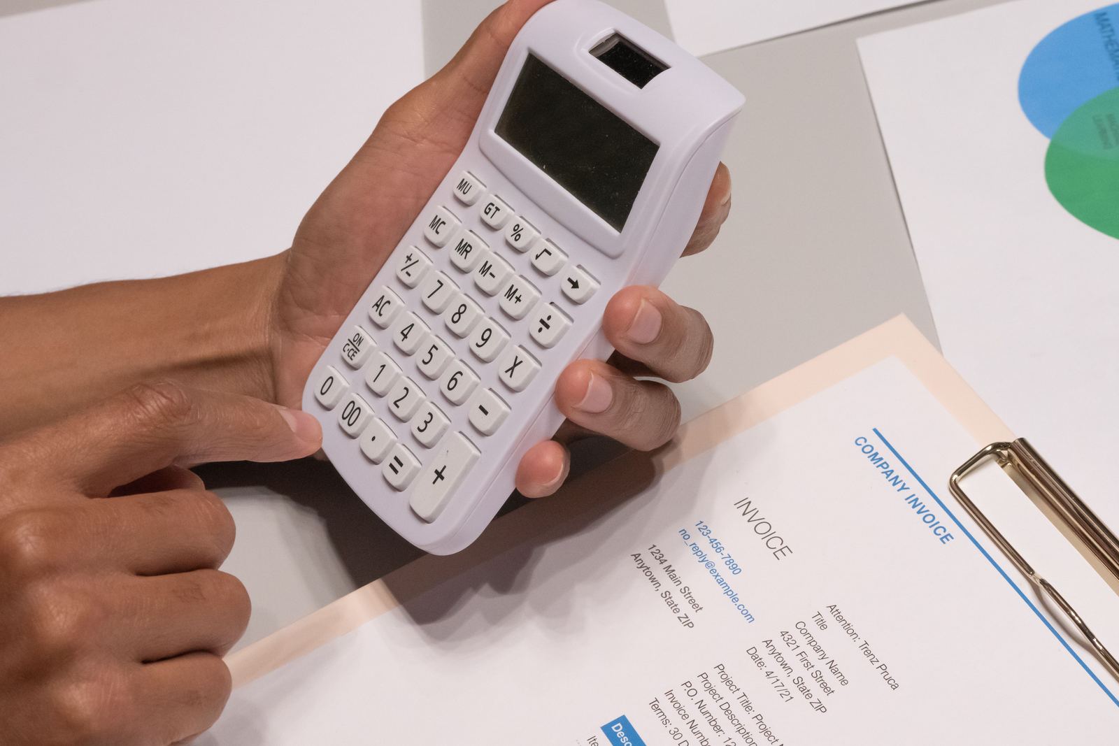 Hands holding a white calculator above a printed invoice on a clipboard
