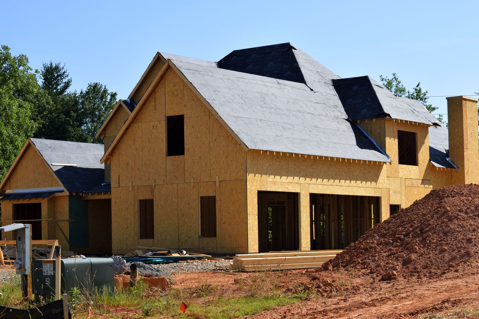 New timber-framed house under construction with boarded walls, dark roof underlay and a pile of soil