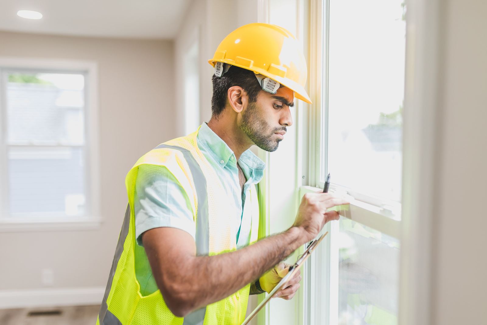 Man in a yellow hard hat and hi-vis vest writing on a clipboard beside a window indoors