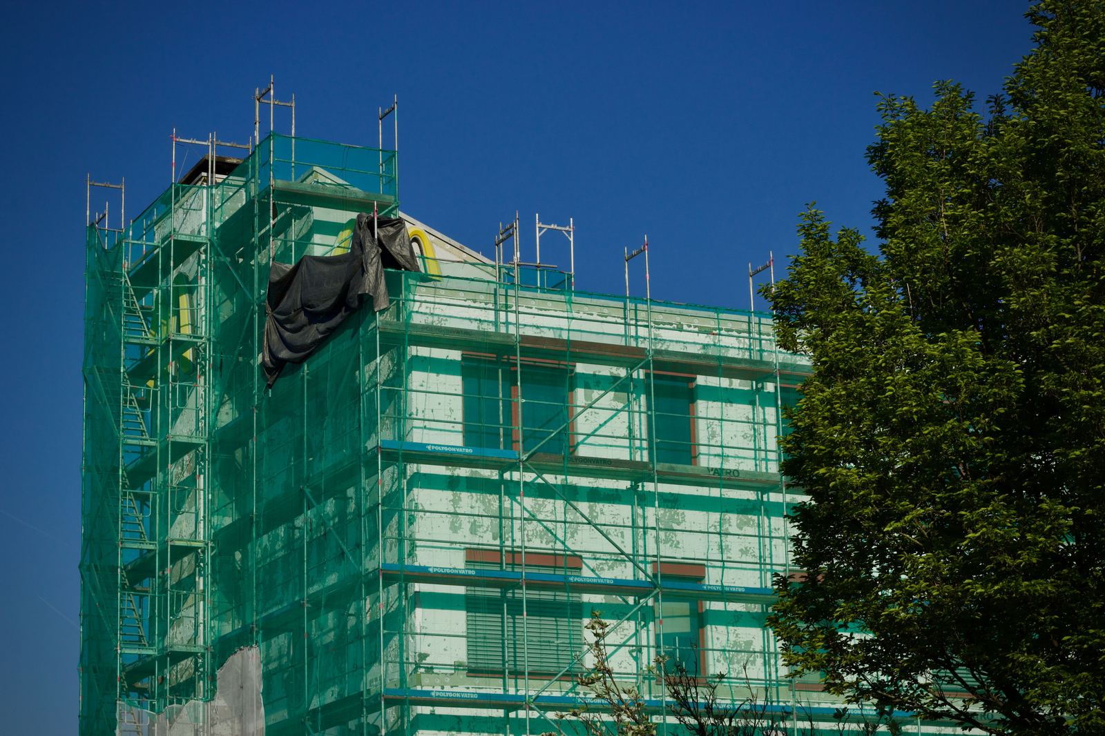 Building wrapped in green scaffold netting with a loose black sheet, beside a tree under blue sky