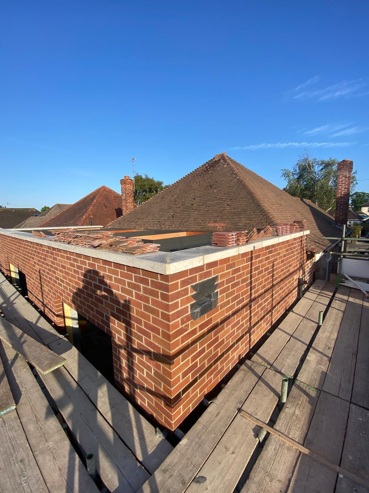 Brick extension walls up to the coping seen from scaffolding, with clay roof tiles stacked on the wall head